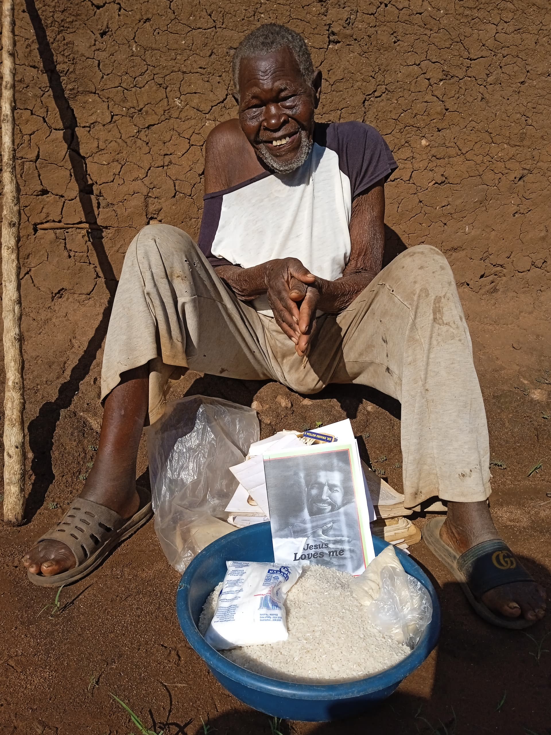 Elderly man Mzei Samuel seated outdoors on a stool, holding a photo card beside a blue basin of rice.