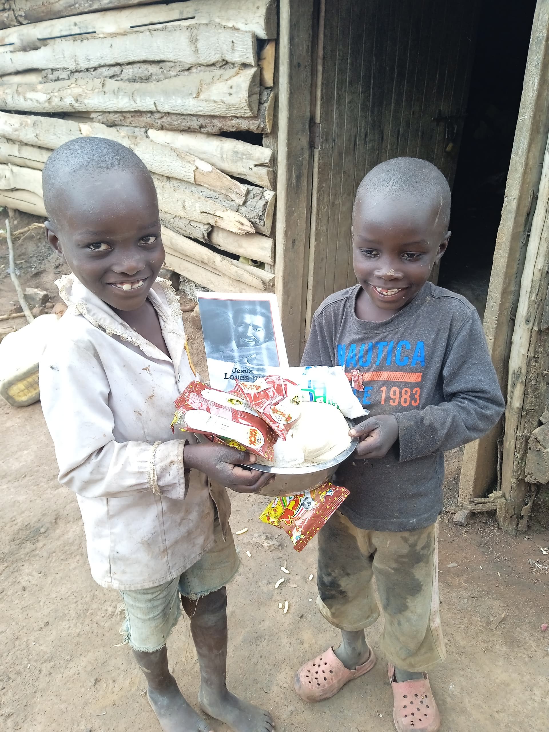 Two children standing outdoors near a wooden hut holding a bowl of food and a photo card.