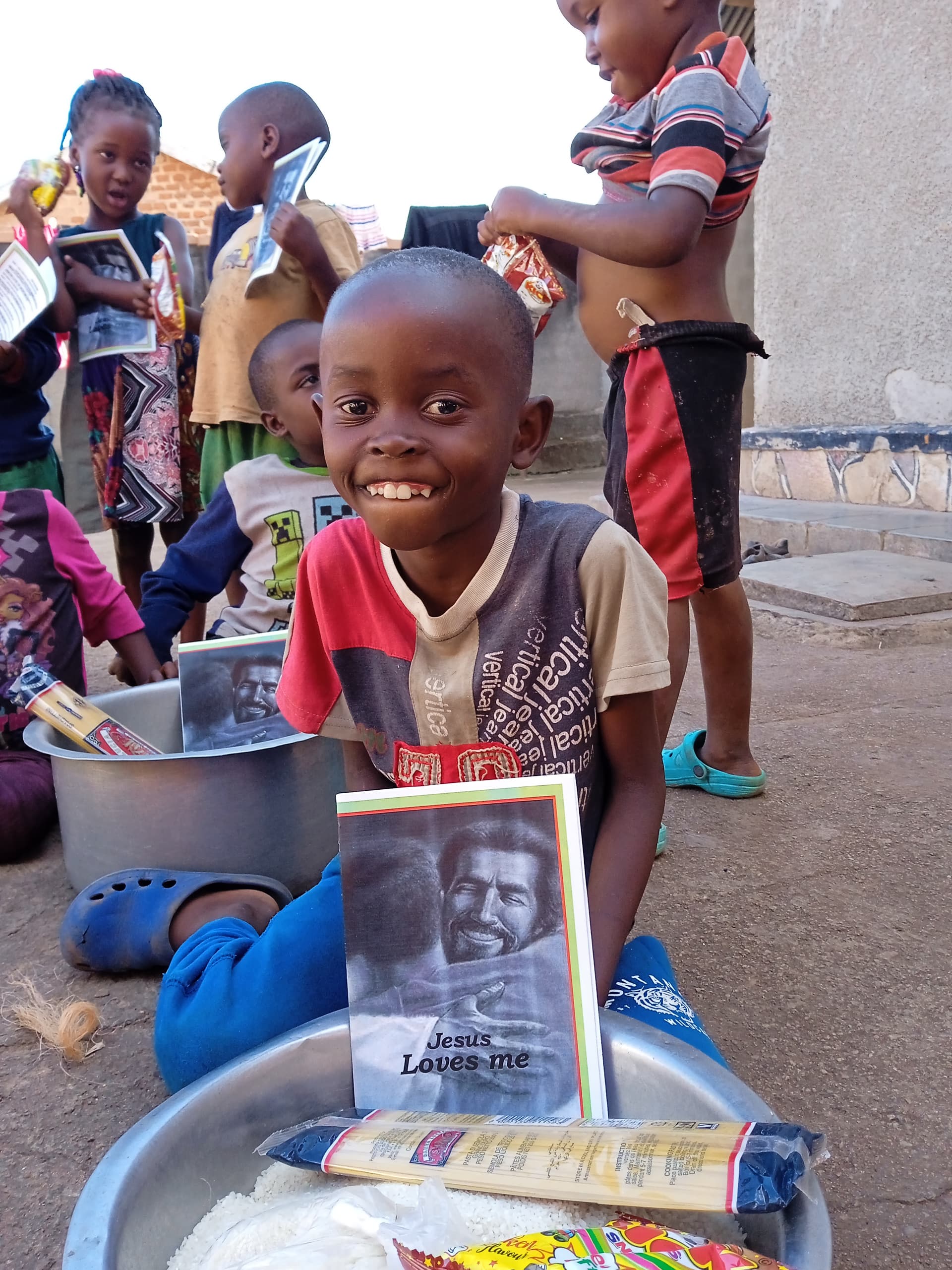 Child sits with a metal basin of food and a photo card while other children gather in the background.