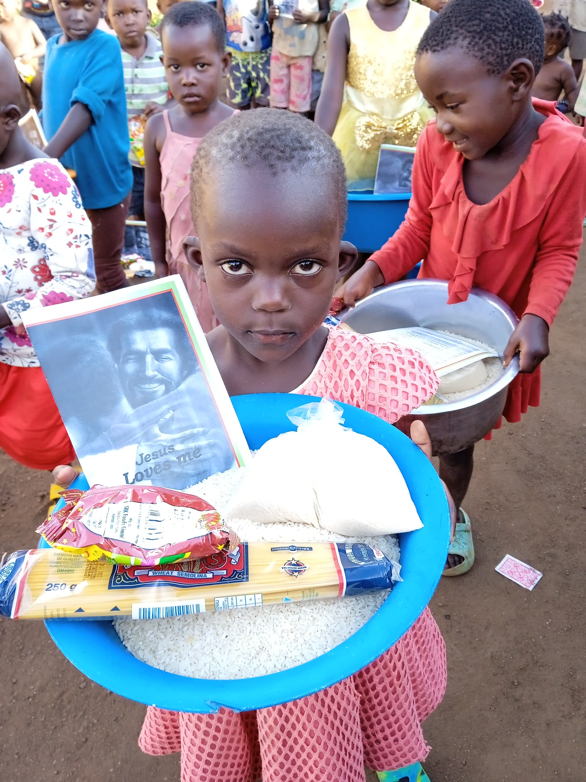 Girl outdoors holding a blue basin filled with rice, a photo card, and other food items, surrounded by a crowd.