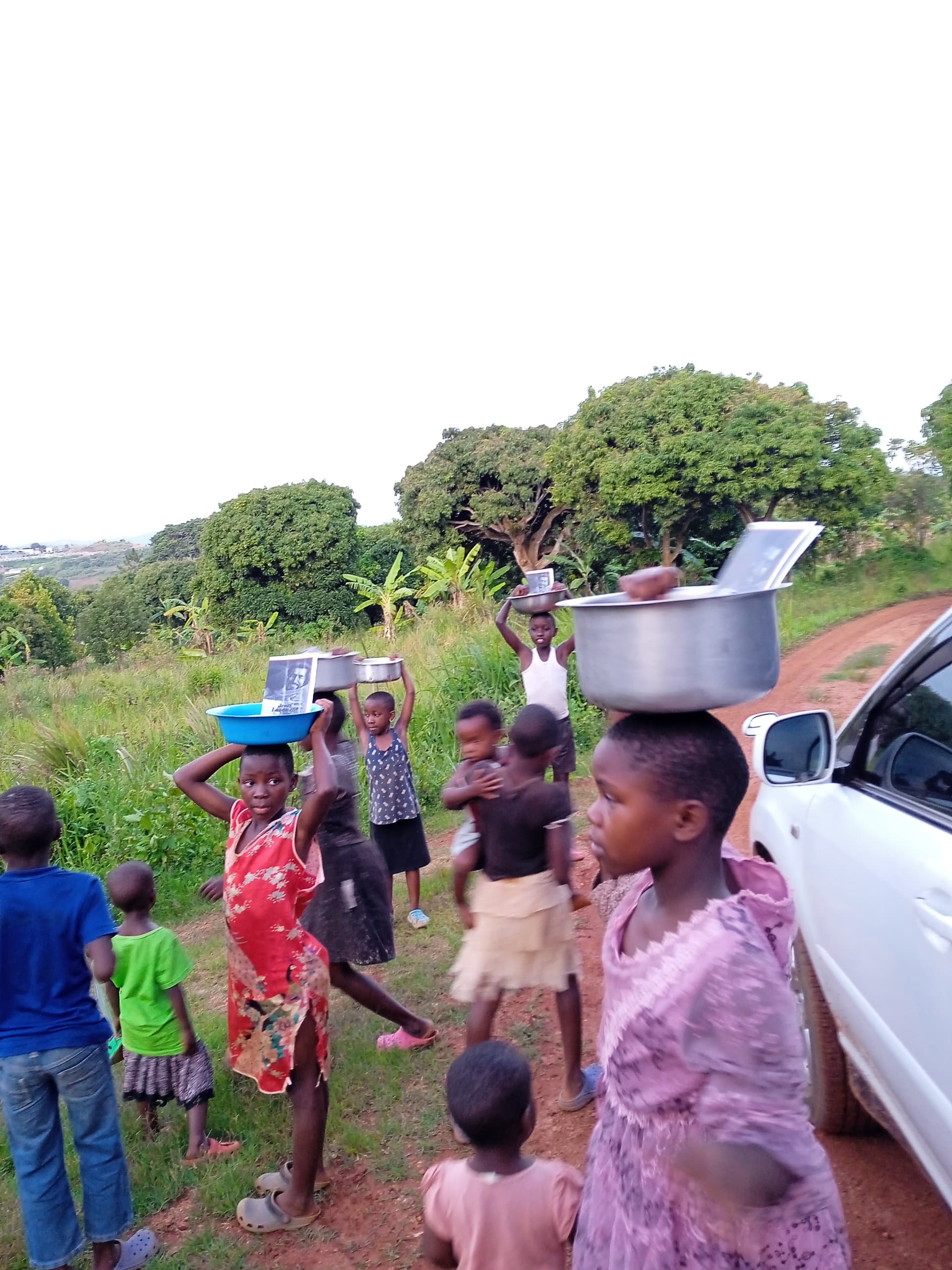 Children walking along a dirt road, one carrying a metal pot on their head, a car parked nearby.