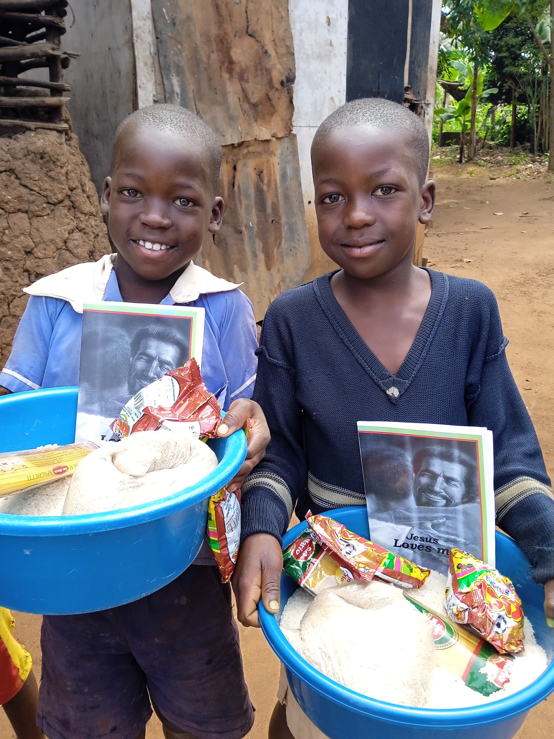 Two boys standing outdoors, one holding a blue basin with a photo card and food.