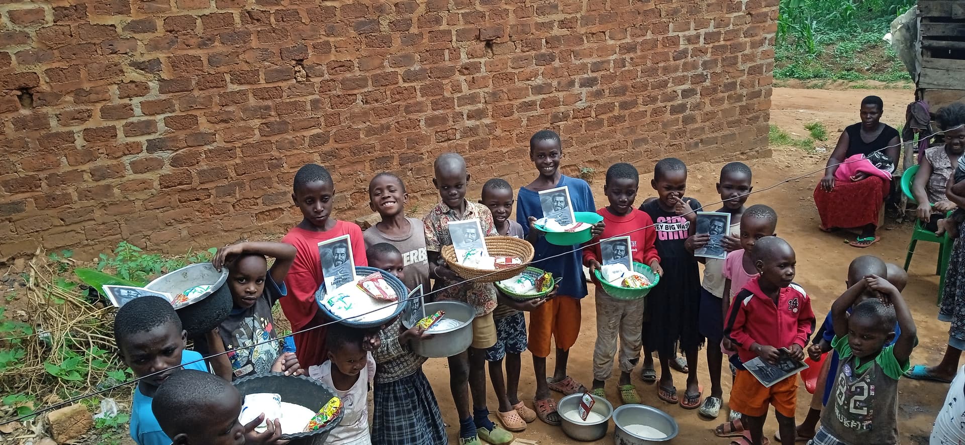 Group of children lined up outdoors against a brick wall, each holding a plate or bowl of food.