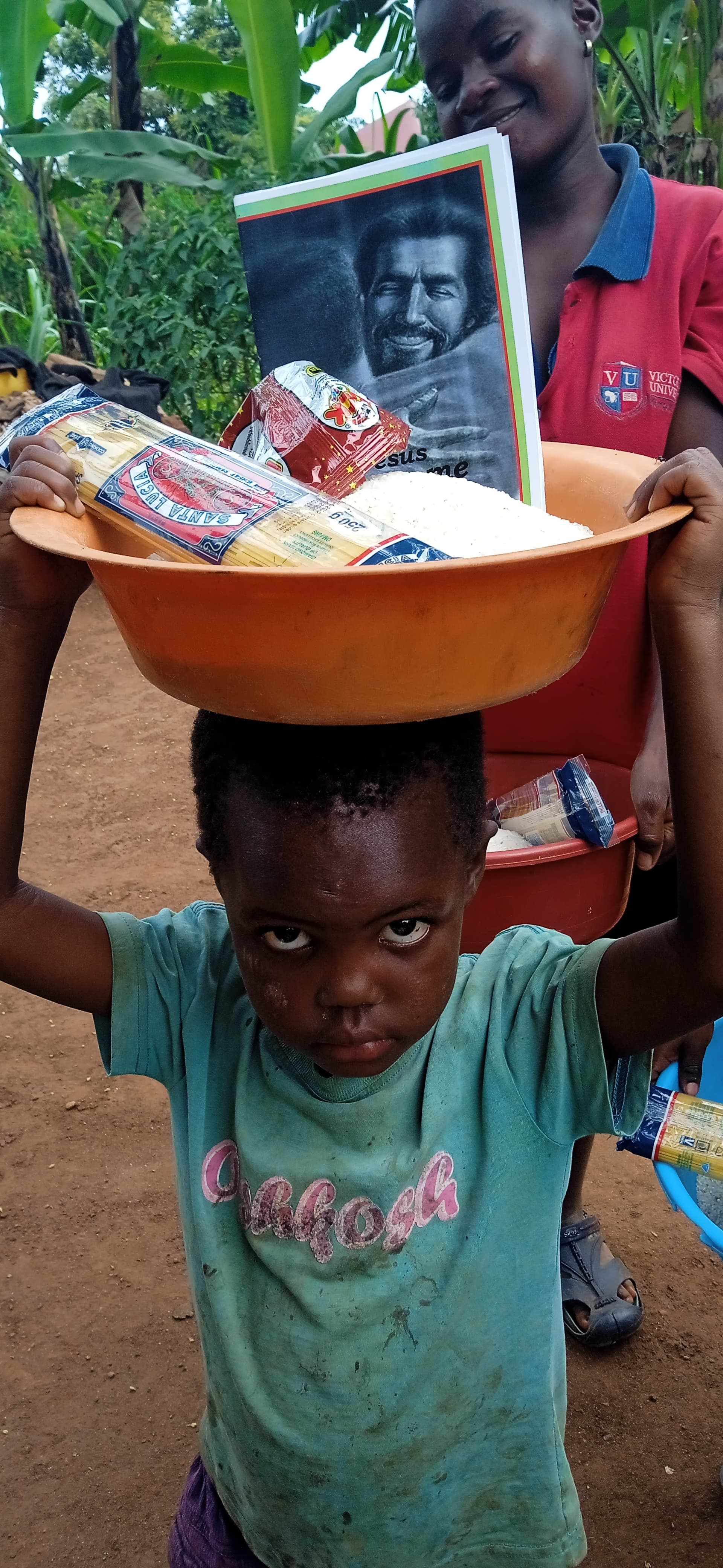 Young boy balancing an orange basin of food supplies and a photo card on his head outdoors.