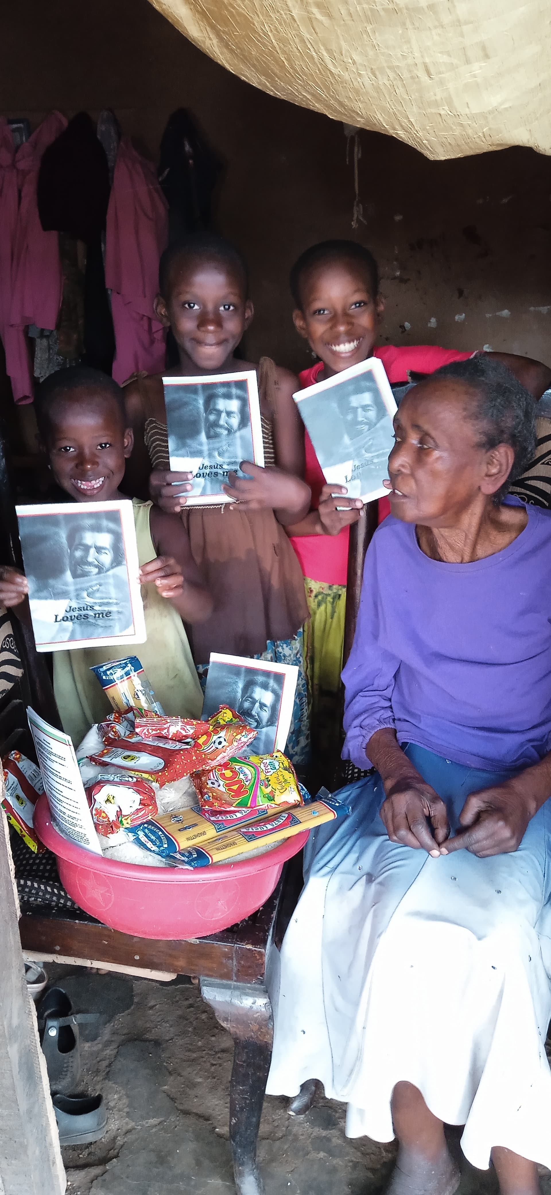 Widow Joeria seated in a purple top indoors under a thatched roof, surrounded by children holding a photo card and food basin.