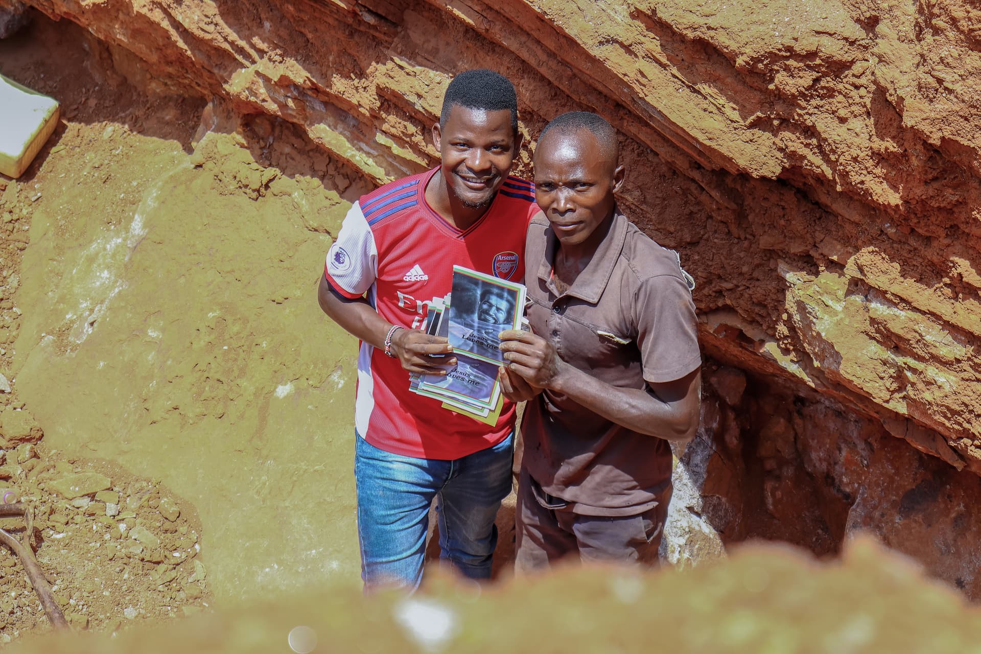 JFM staff member in a red shirt stands with a quarry worker, both holding a printed gospel booklet.
