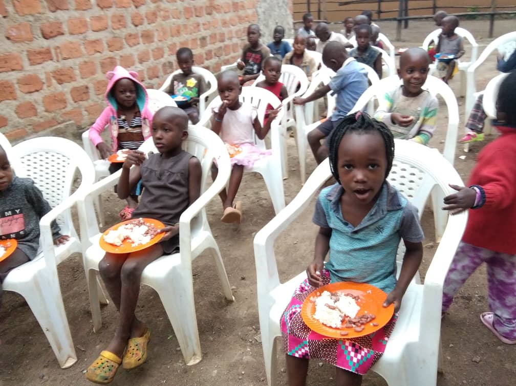 Children sit in white plastic chairs against a brick wall eating food from plates.