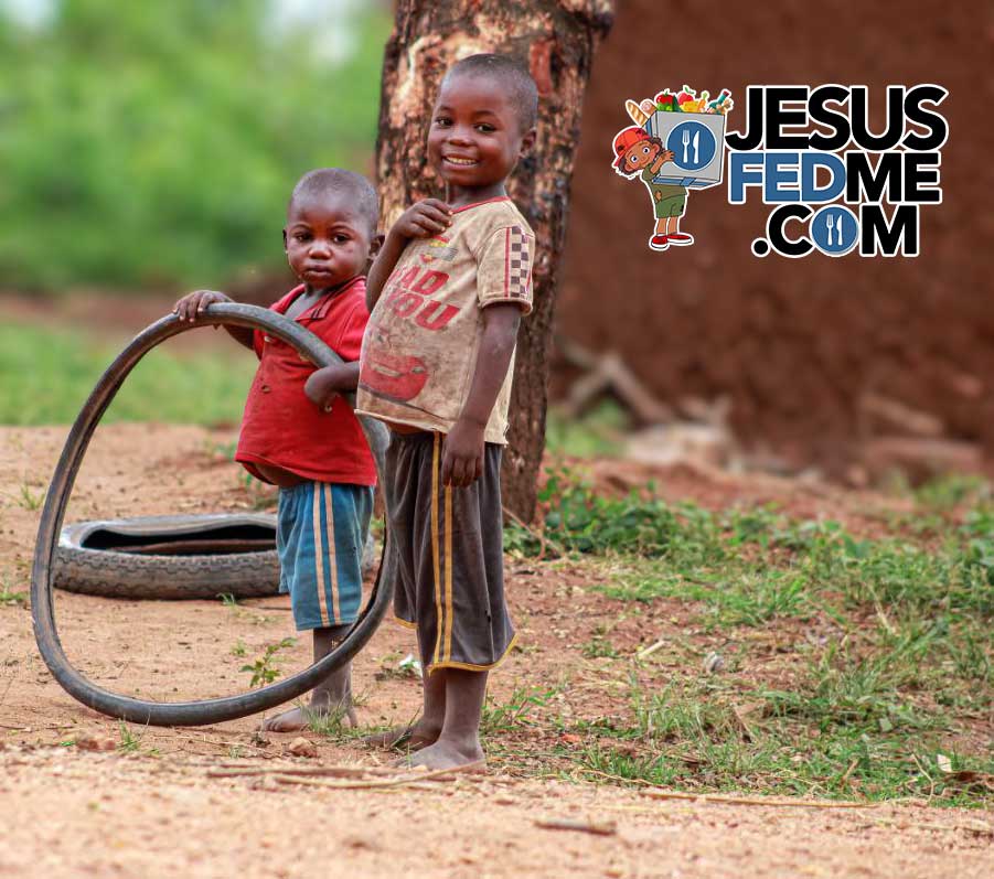Two children stand beside an old tire on a dirt path, one holding a stick.