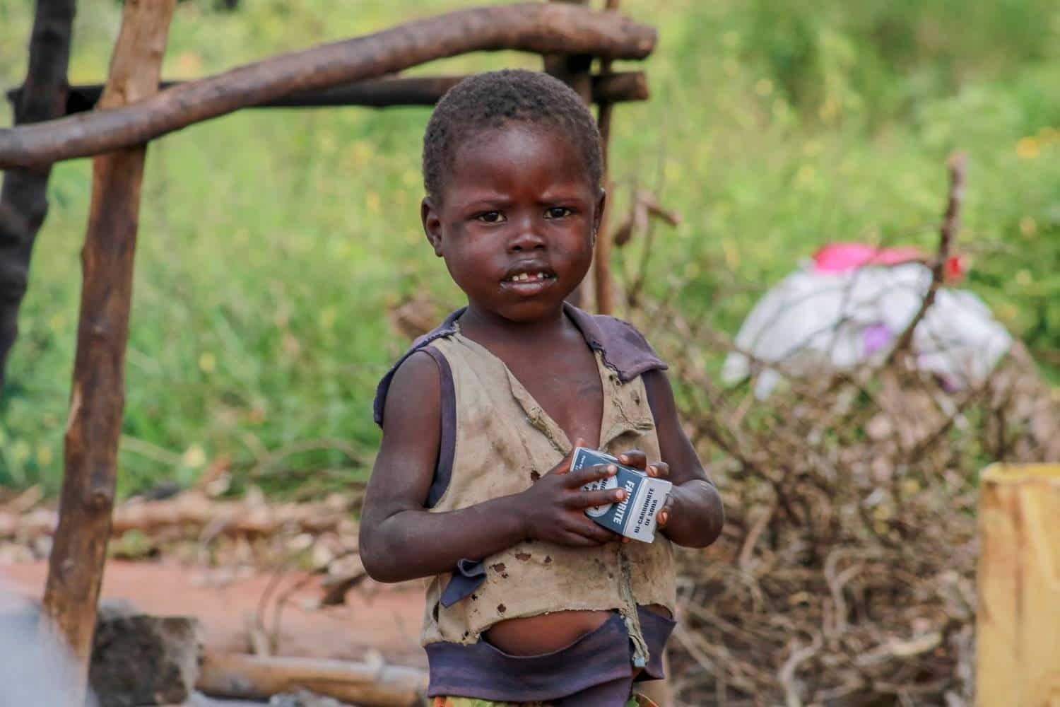A young child in a worn shirt stands outdoors near a wooden structure holding a small item.