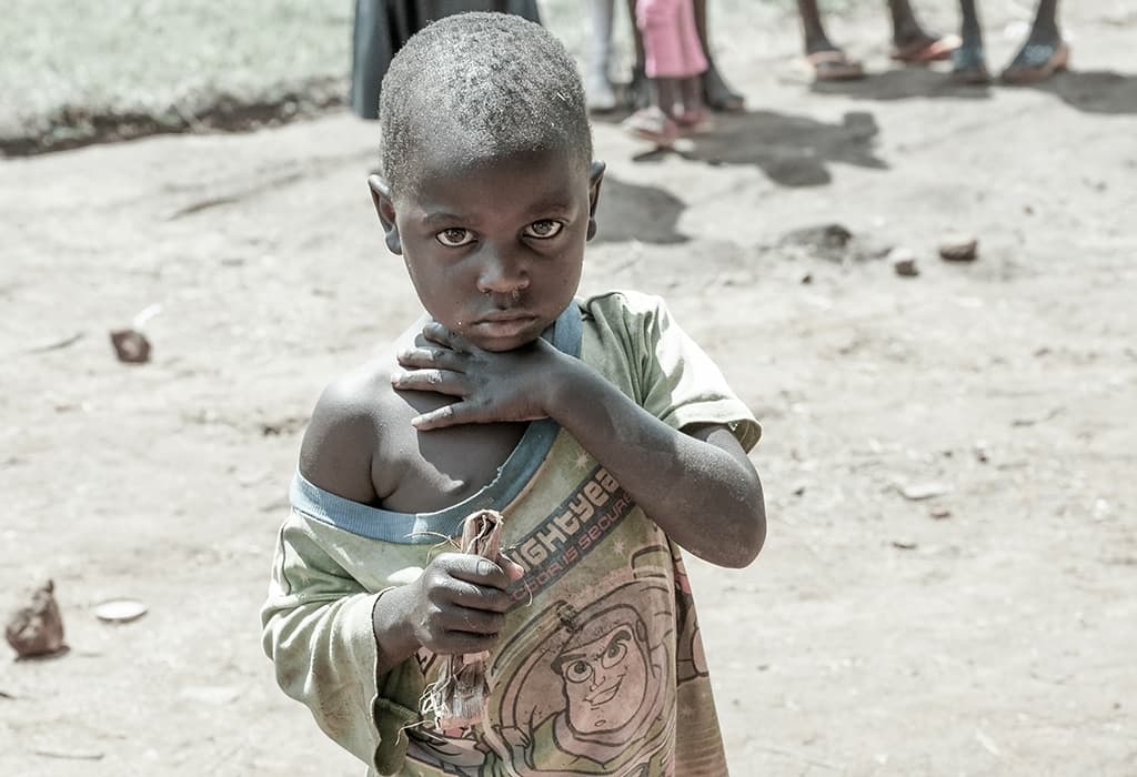 Young boy stands on dirt ground with his shirt pulled up, arms crossed over his bare chest.