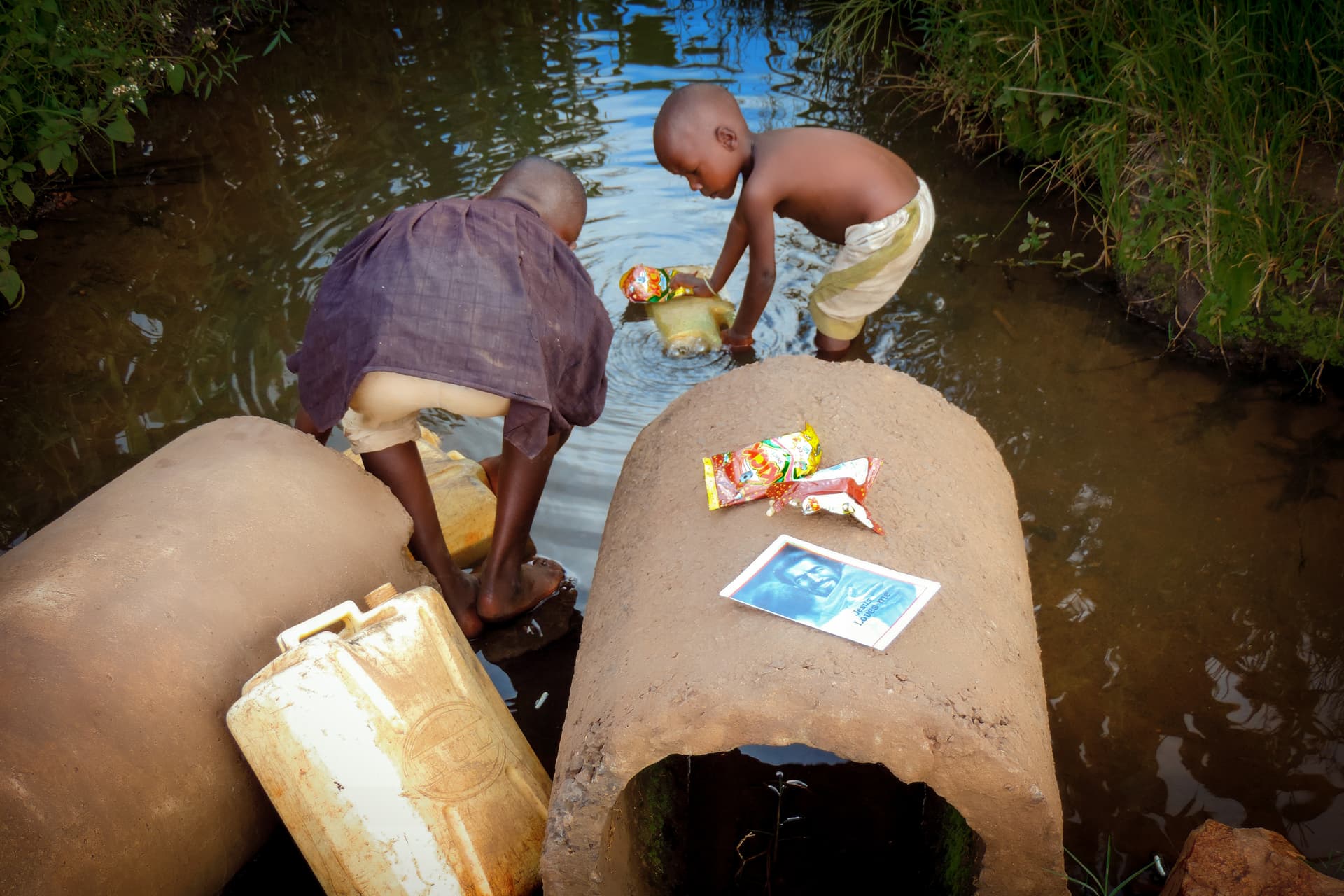 People fill large yellow jerry cans with water at a stream, a gospel tract resting on one can.