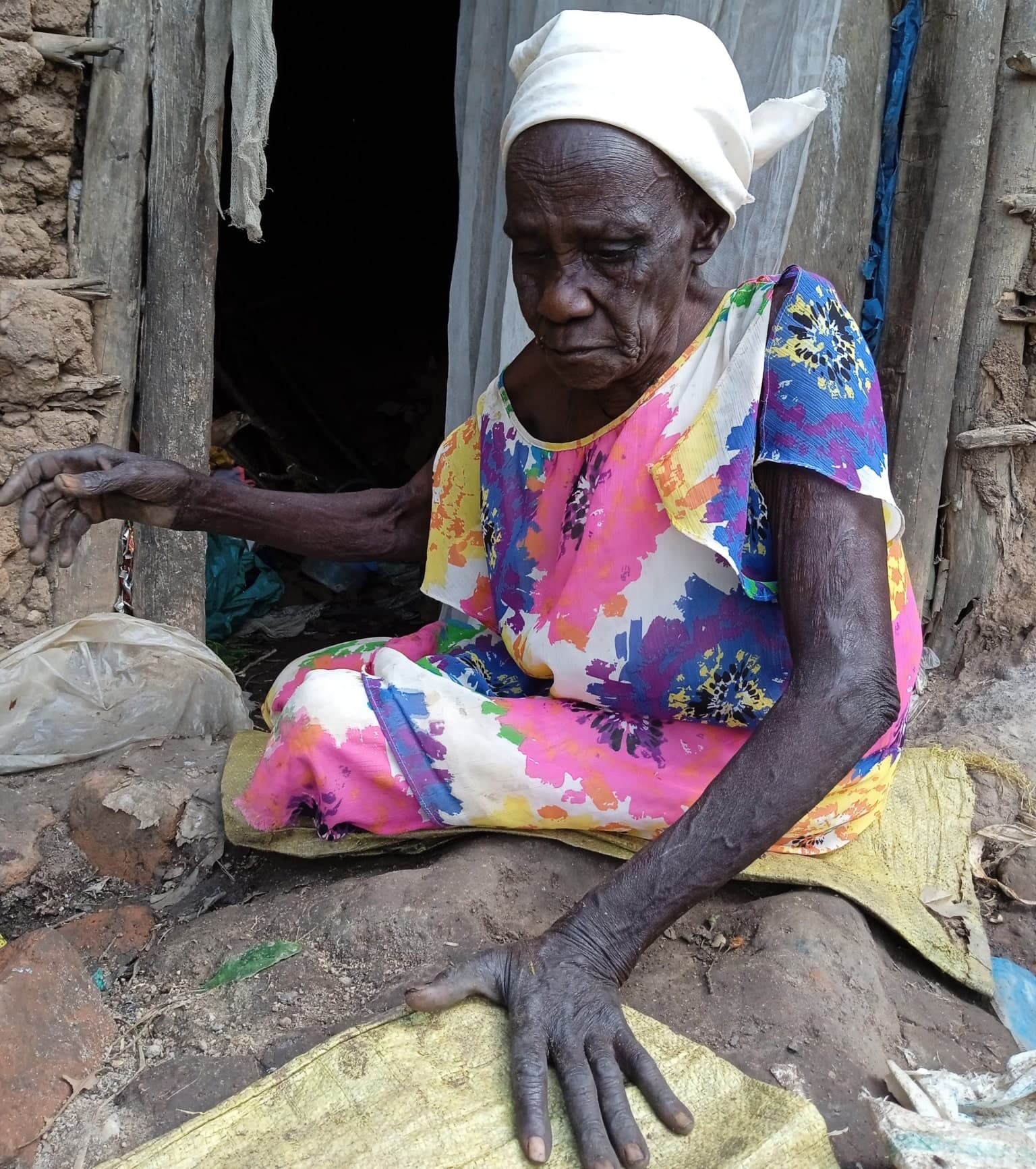 Elderly woman in a headscarf and floral dress sits on the ground outside a mud-brick home, reaching out a hand.