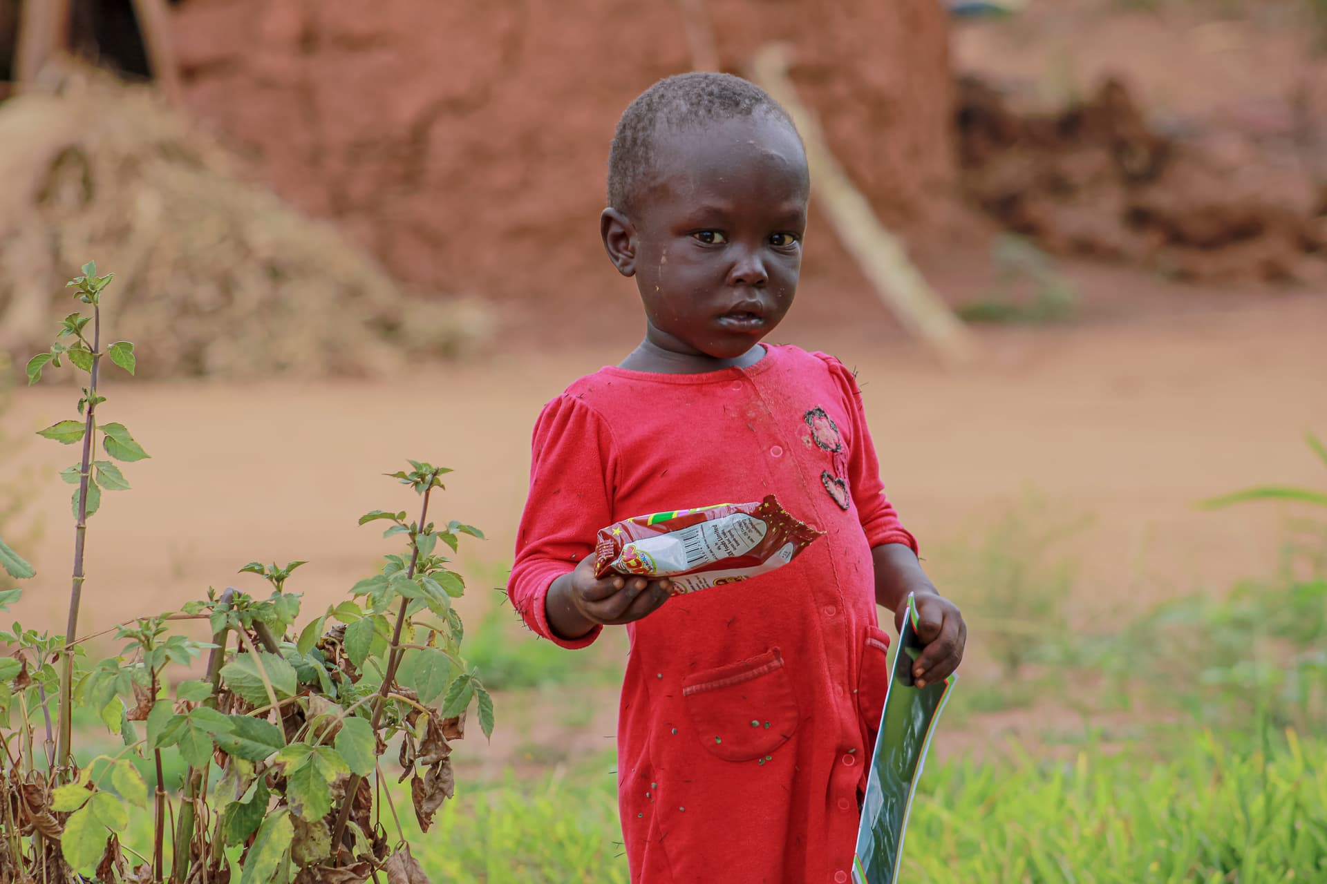 A young child in a red shirt stands outdoors on a dirt path holding a small item.