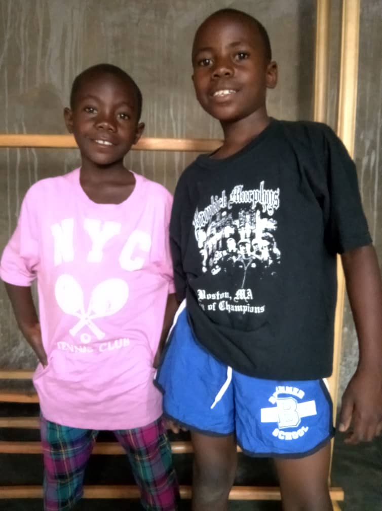 Two boys stand together indoors in new outfits, one in a pink graphic shirt, the other in a dark tee and blue shorts.