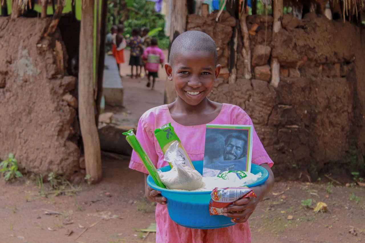 A smiling girl carries a blue basin of food supplies and a gospel tract along a village path.