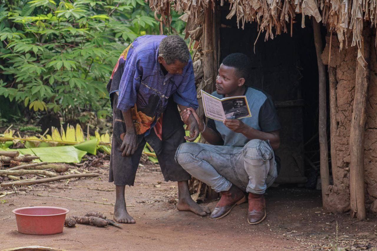 An elderly man reads from a book to a young man seated outside a mud-brick home, a red basin nearby.