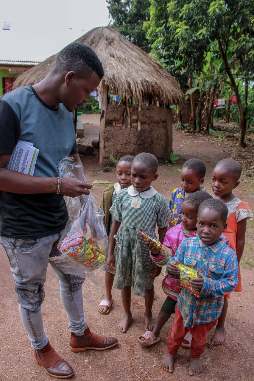 A young man in a cap hands out food bags to a group of children outside a thatched hut.