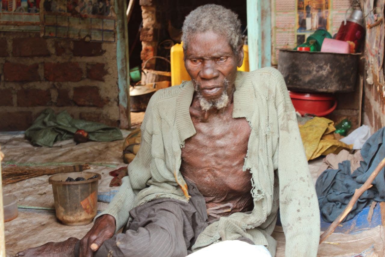 Frail, malnourished elderly man sitting on the floor inside a bare brick room.