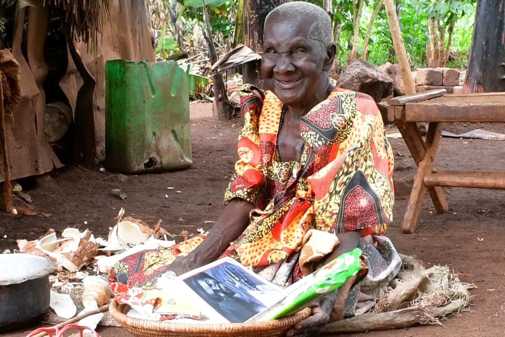 Elderly woman seated near banana plants, smiling, with a food package and cooking pot beside her.
