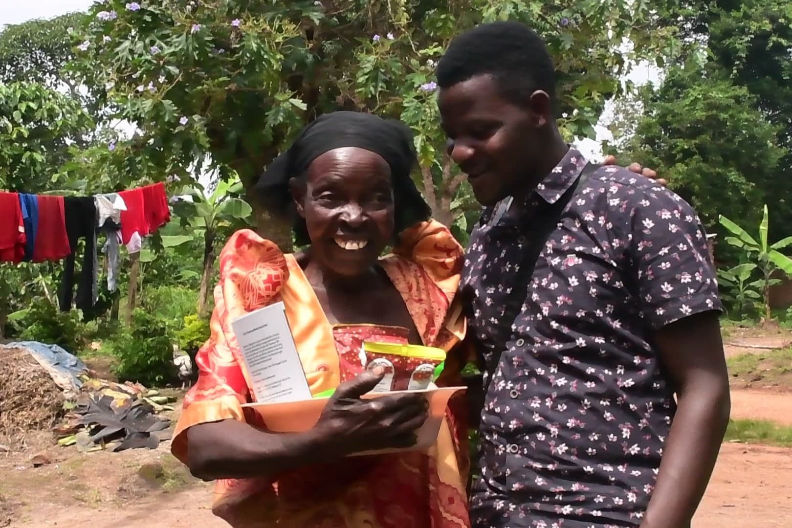 A woman and a young man stand together outdoors smiling, holding a booklet, with laundry hanging on a line behind them.