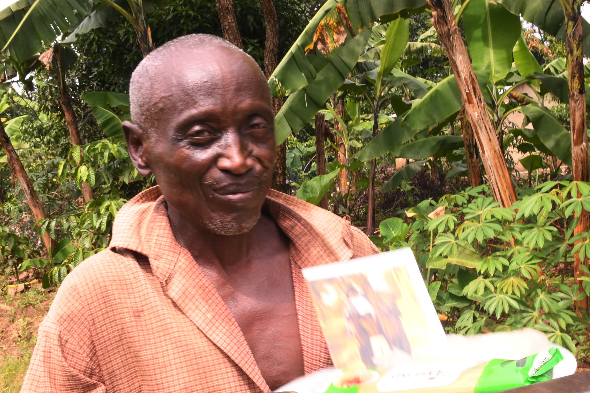 An elderly man stands among banana plants holding a food gift package wrapped in bright paper.