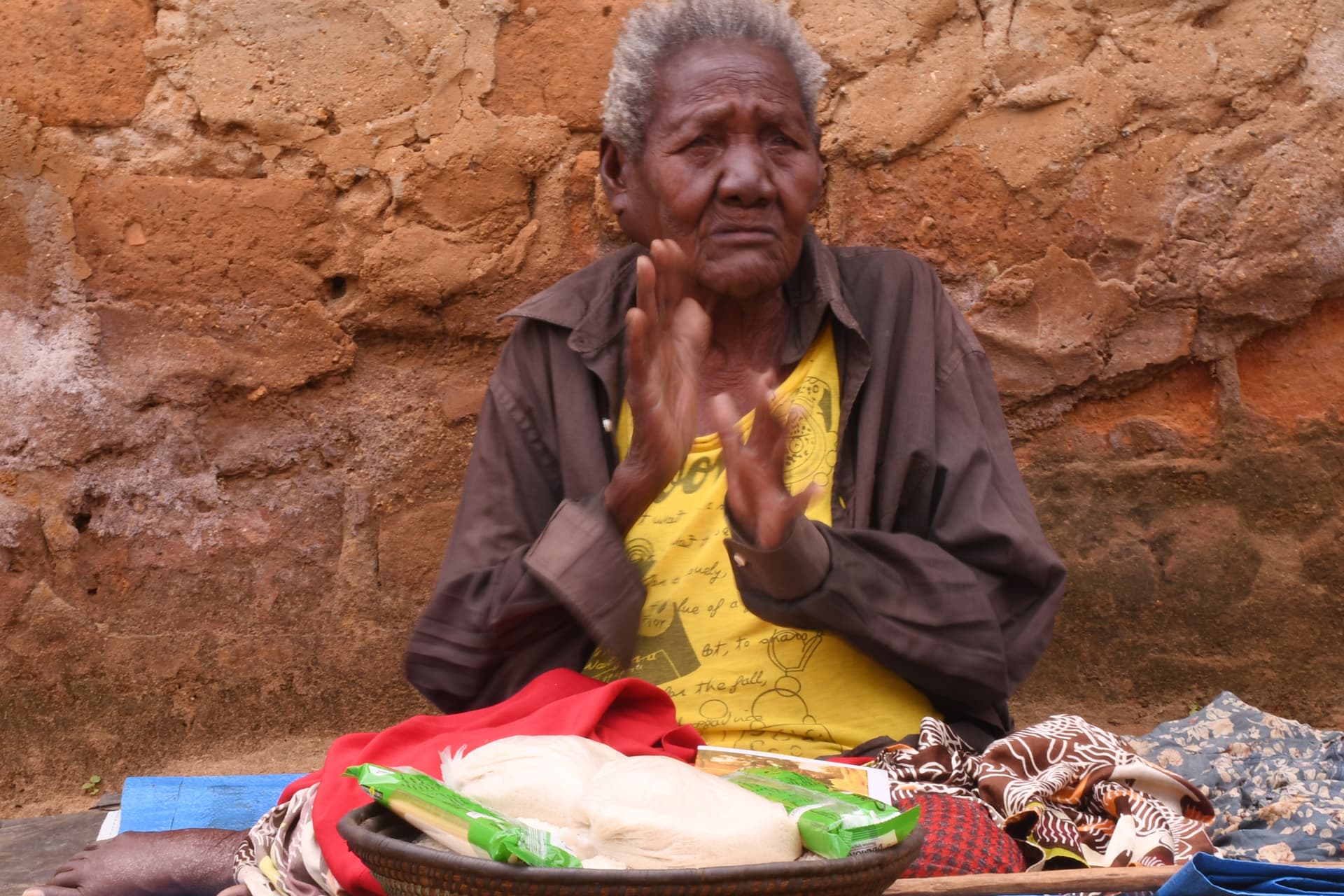 An elderly woman sits on the ground with hands clasped in thanks, food and gift items spread on a cloth before her.