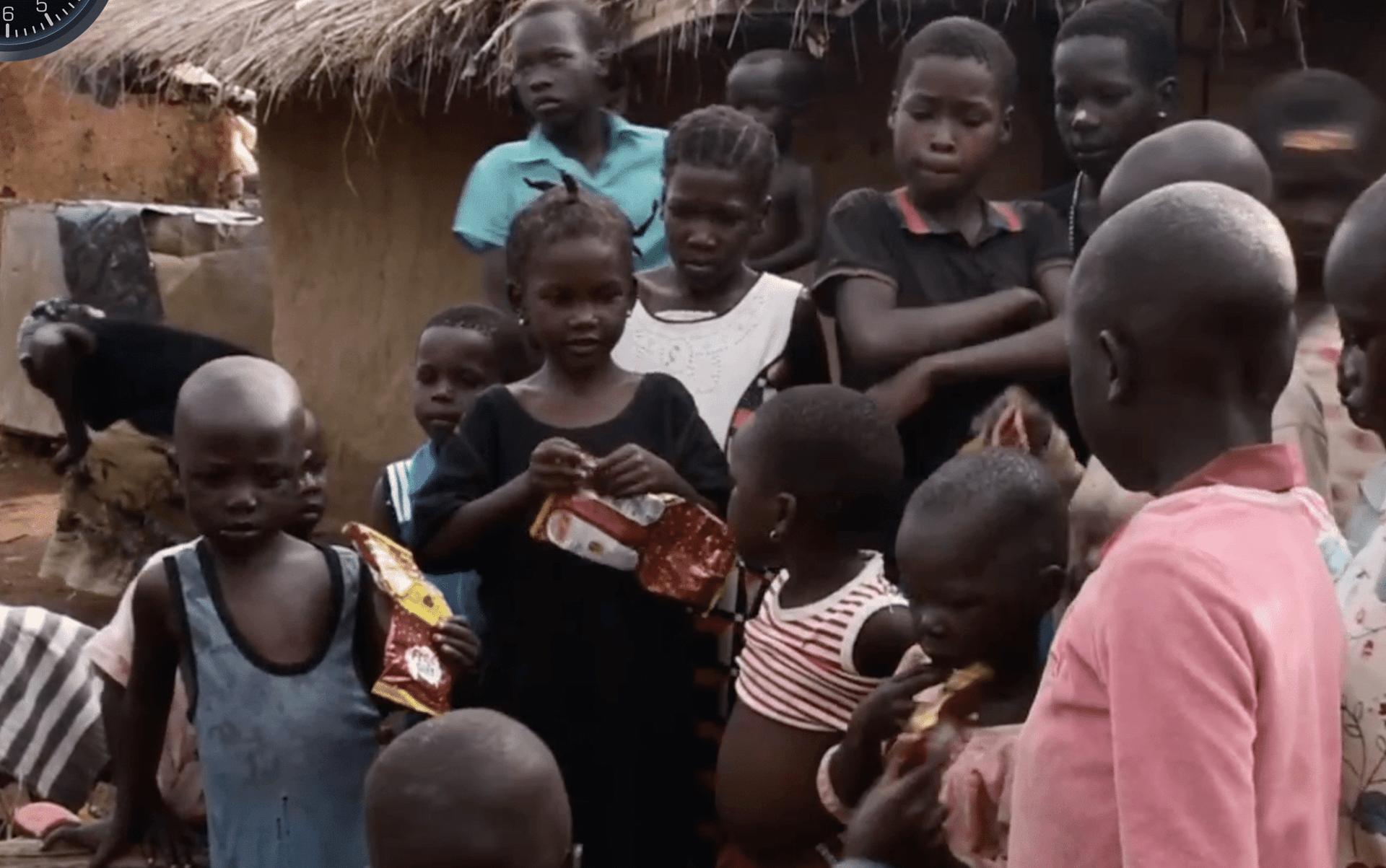 A crowd of children gathers outside a thatched-roof hut, some holding gift items and photographs during a Christmas celebration.