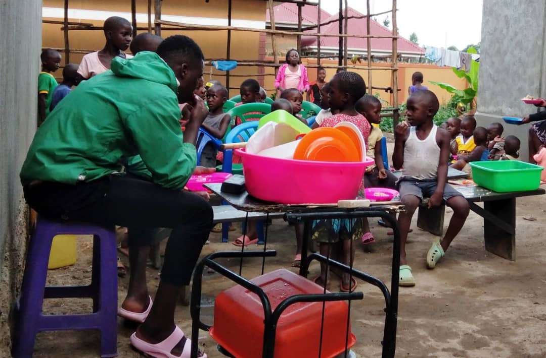 JFM staff member in a green shirt distributing food and toys to children gathered at a gated compound.