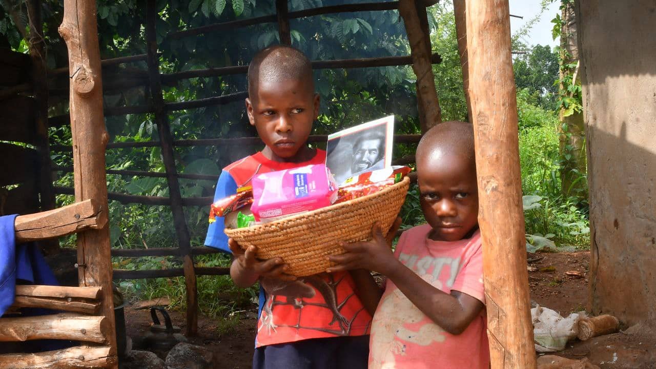 Two children standing under a wooden shelter holding a basket with a food package.