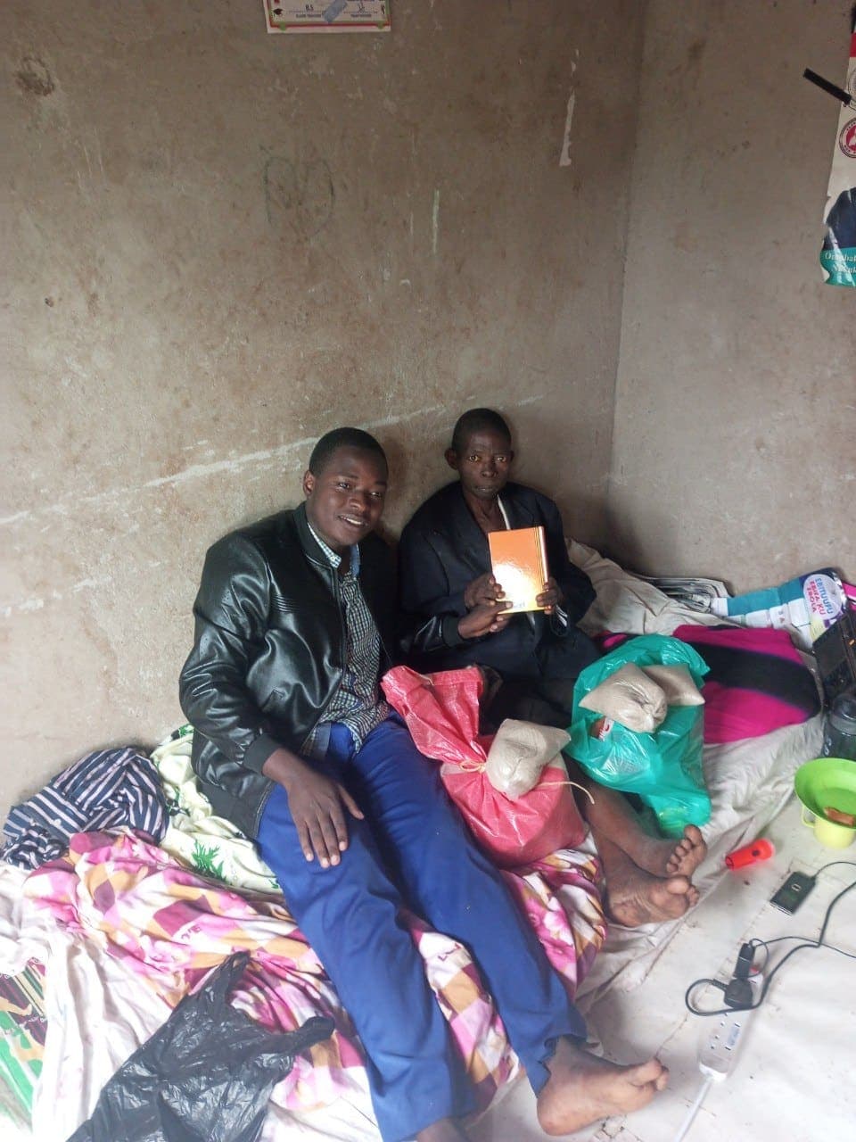 Two men sitting together on a bed in a sparsely furnished room, one holding a document.