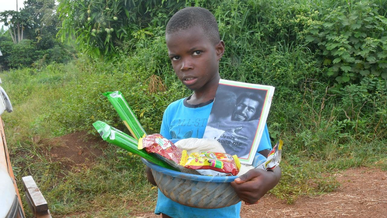 Boy standing on a dirt path holding a basket with a food package and green produce.