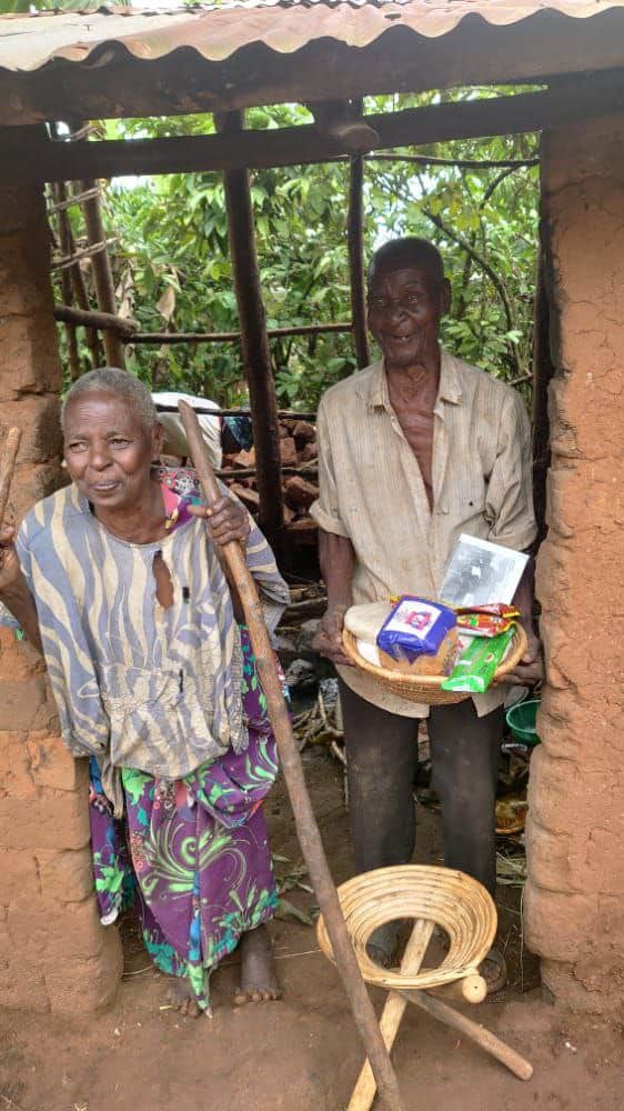 An elderly couple stands outside their home holding a food gift package, with a woven basket nearby.