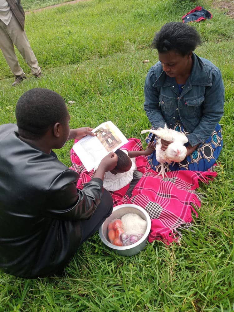 A man and a young woman sit together on the grass sharing a meal from a bowl.