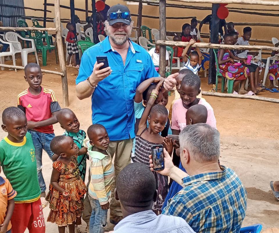 Man in a blue shirt holding a phone, surrounded by children outdoors near plastic chairs.