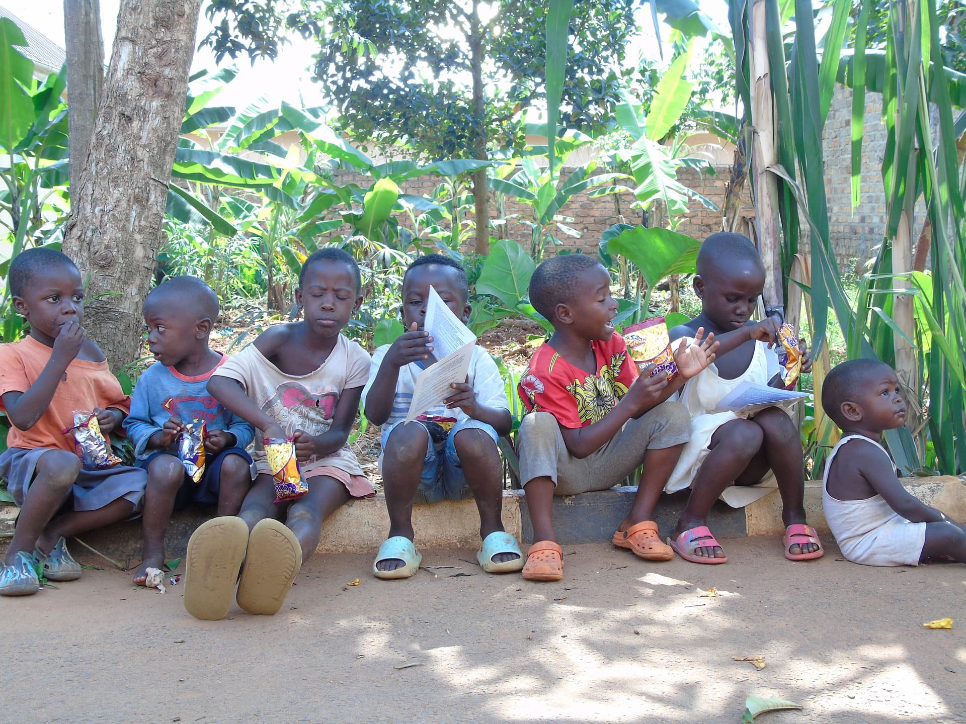 Group of children sitting together outdoors among banana plants, being photographed for a camera test.