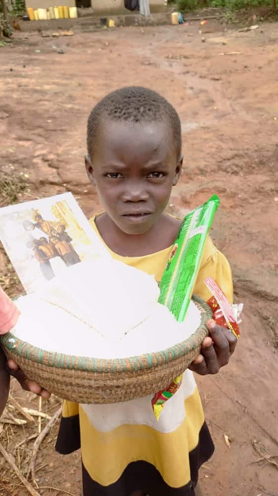 A young boy stands on a dirt path holding a white basin loaded with rice and gift items.