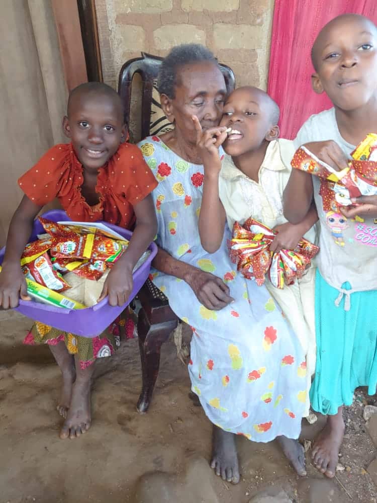 Granny Joeria poses indoors with three young children in patterned clothing, one holding a bag of gift items.