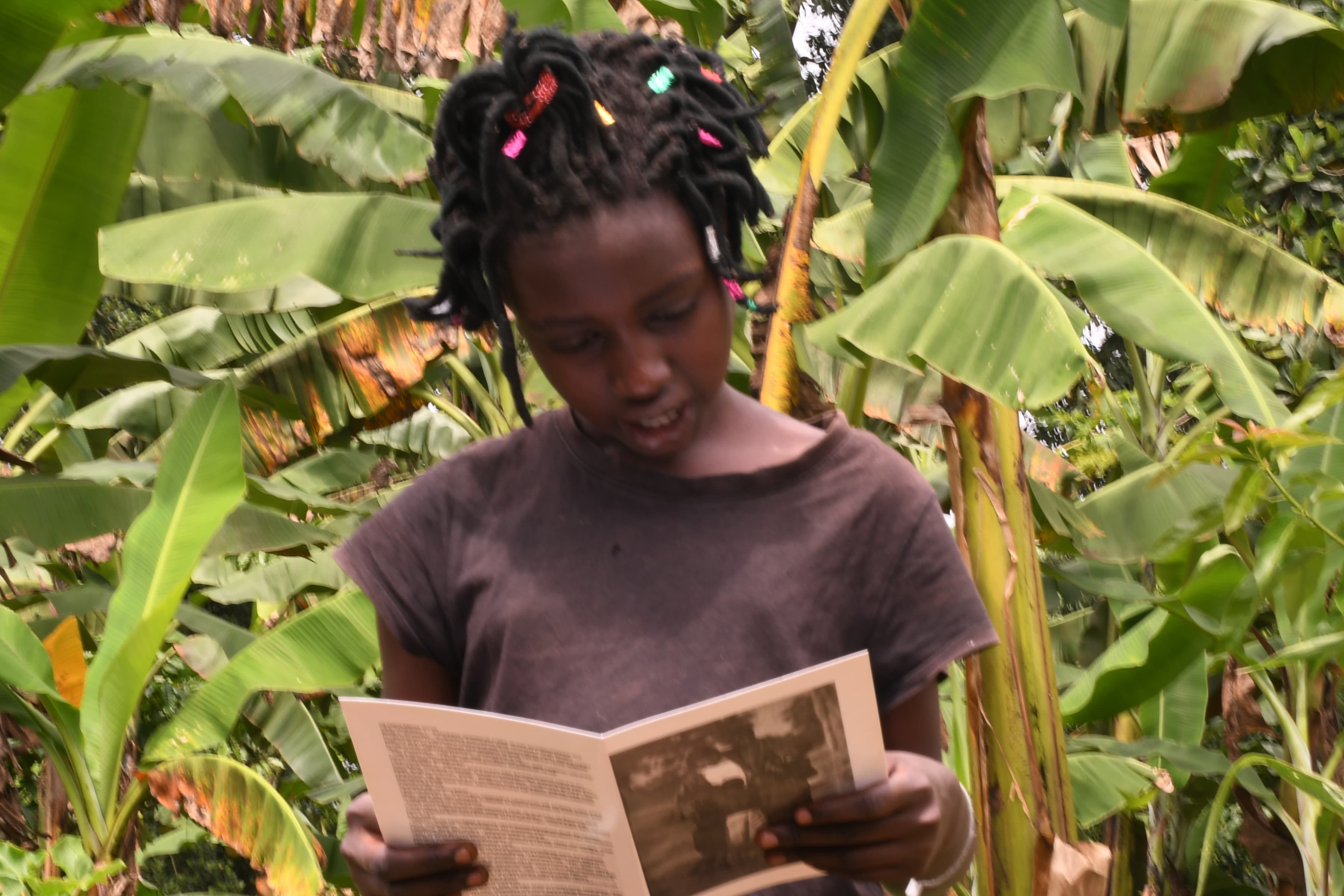 A girl with braided hair reads a pamphlet while standing among banana leaves.
