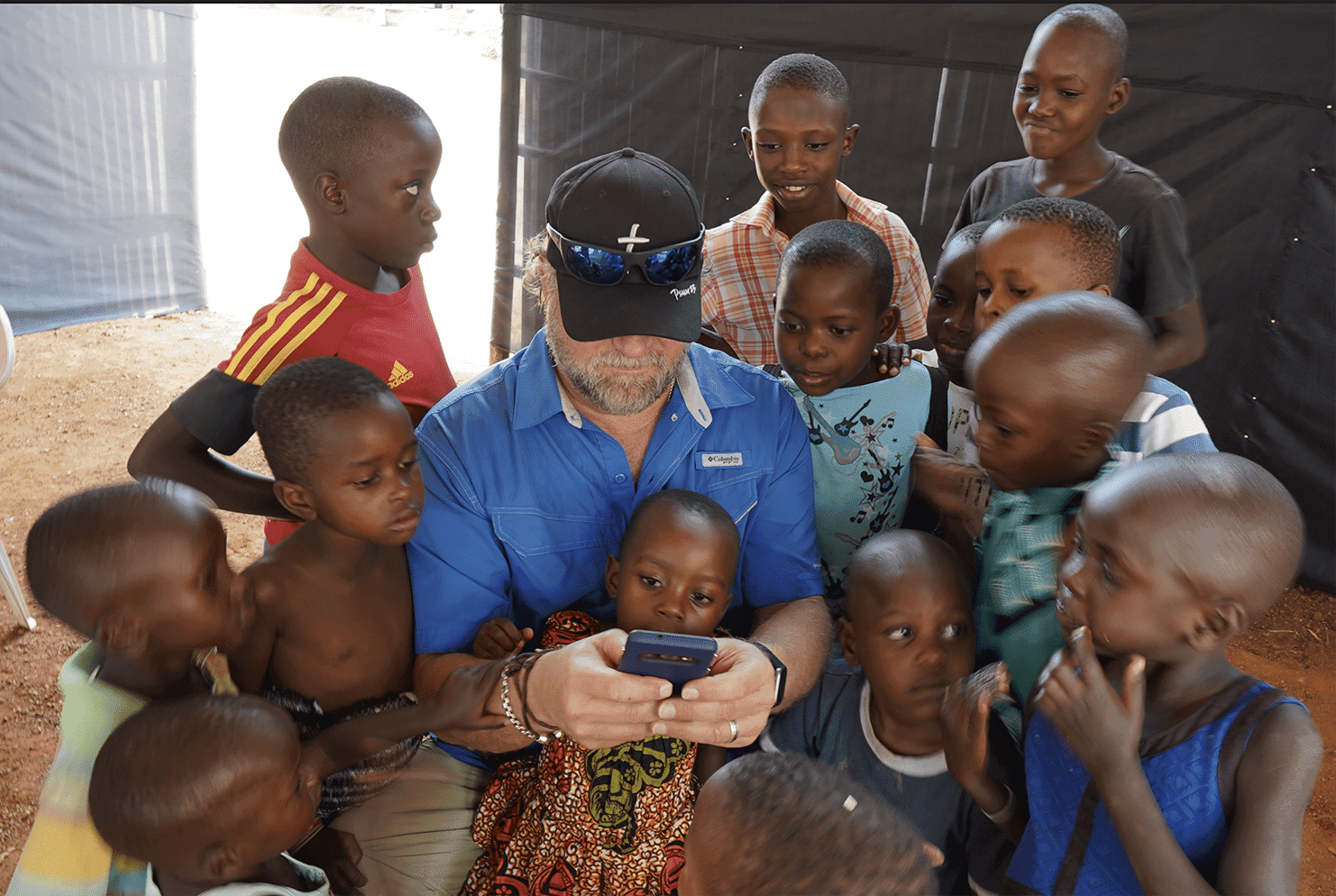 A visitor in a blue cap is surrounded closely by a group of young children at the orphanage.