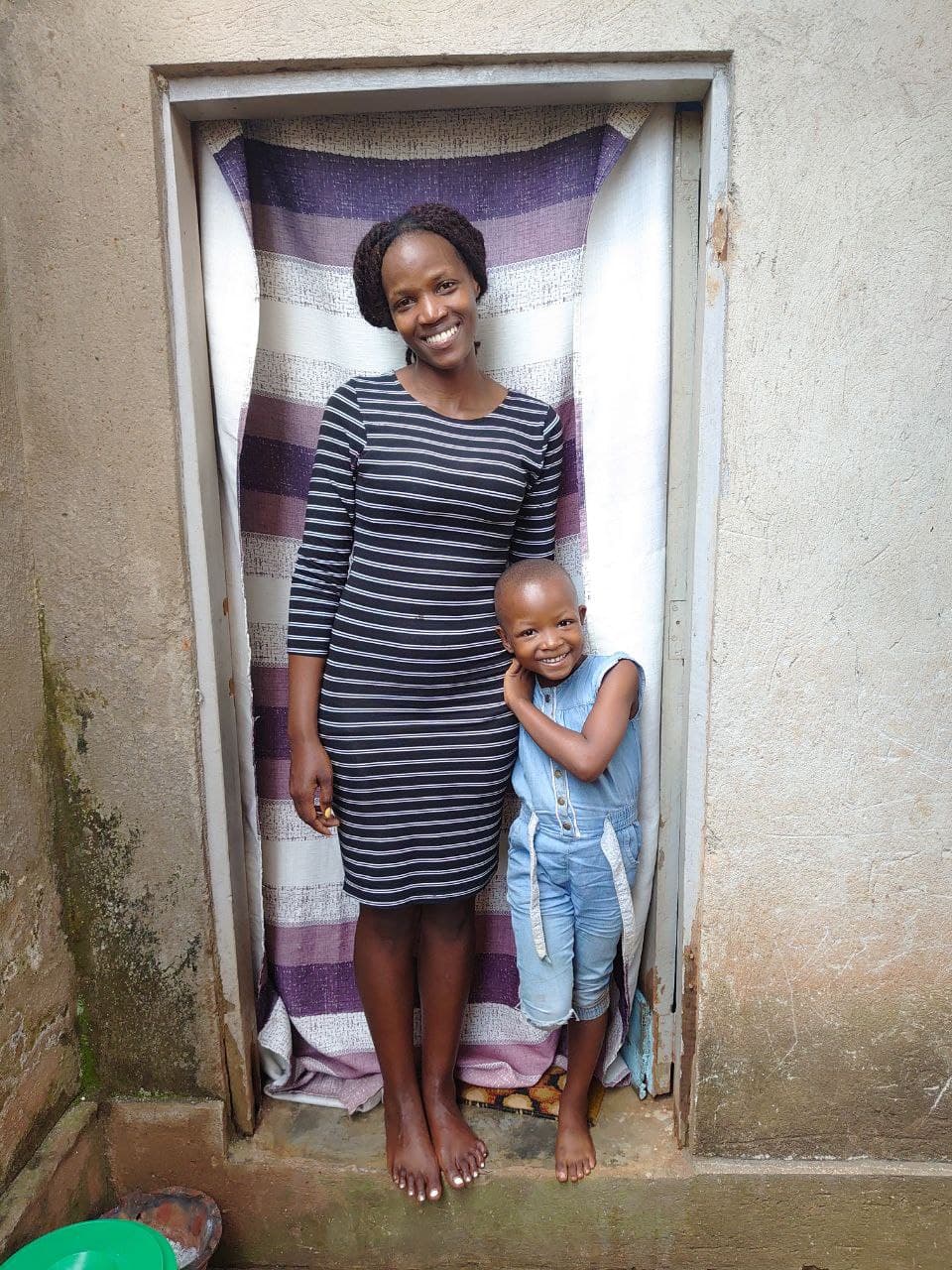 Daisy stands in a doorway with a young girl beside her, striped fabric hanging as a backdrop.