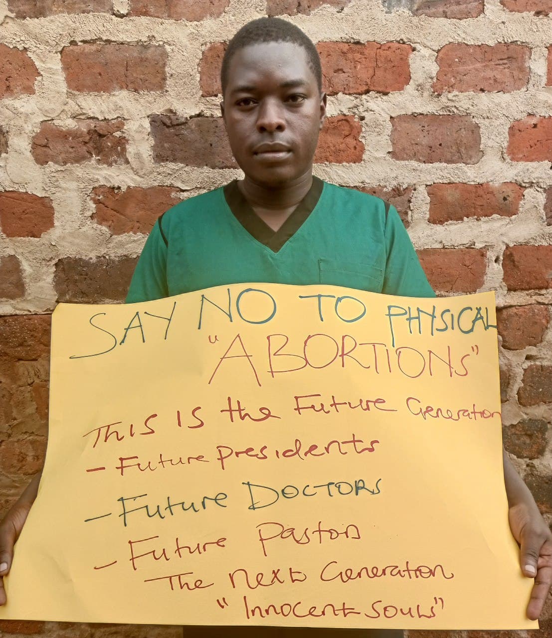 Young man in green scrubs holding a handwritten sign reading 'Say No to Physical Abortions' against a brick wall.