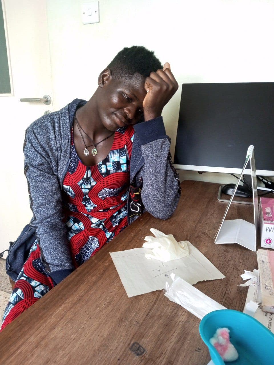 Young woman in a floral dress sitting at a table in a clinic office, head resting on her hand.