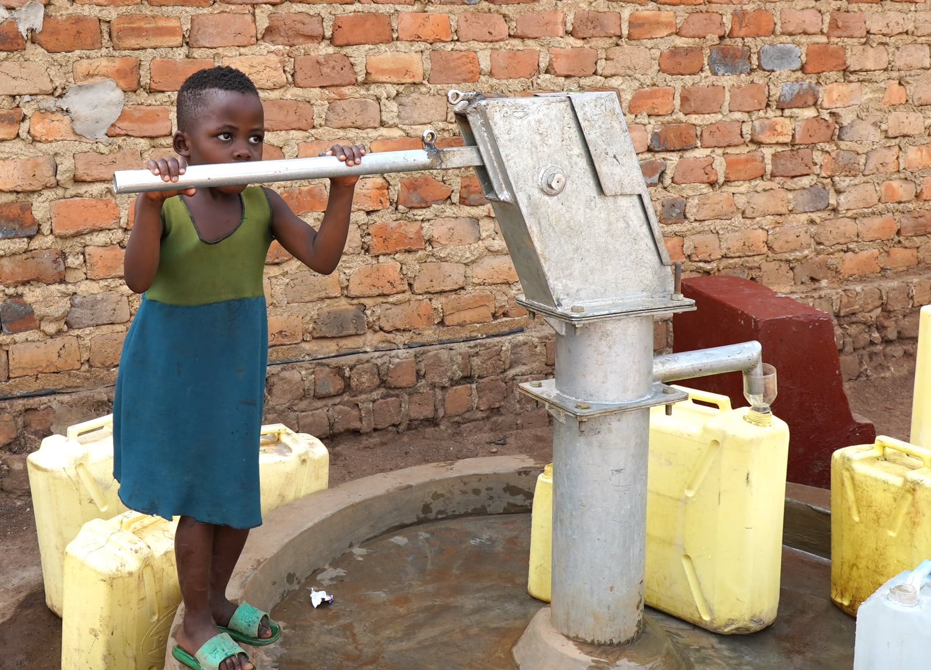 Girl pumping water from a hand-pump borehole into a puddle, yellow jerrycans waiting to be filled around her.