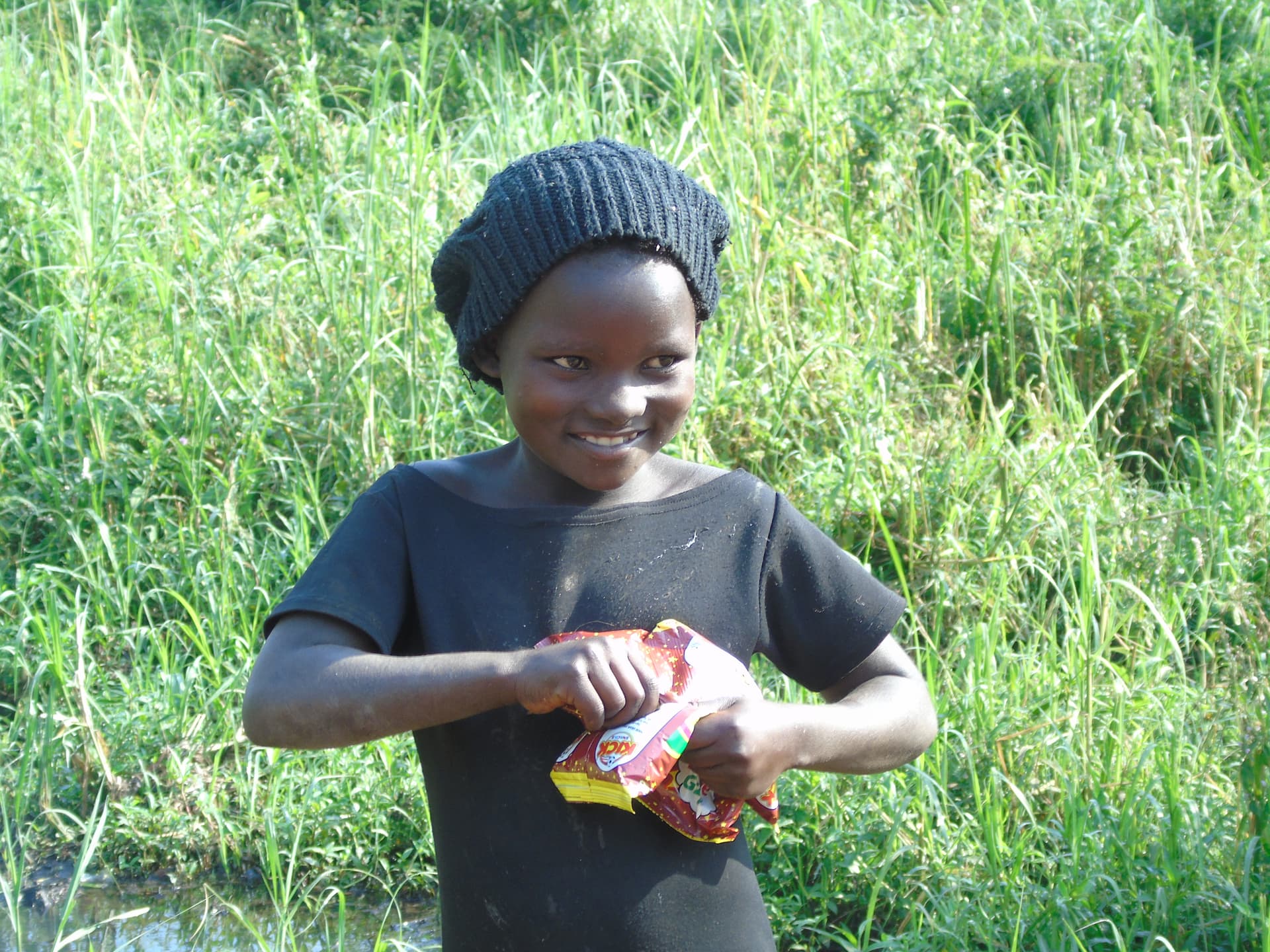 Smiling boy in a gray beanie standing outdoors holding food packets from a relief distribution.
