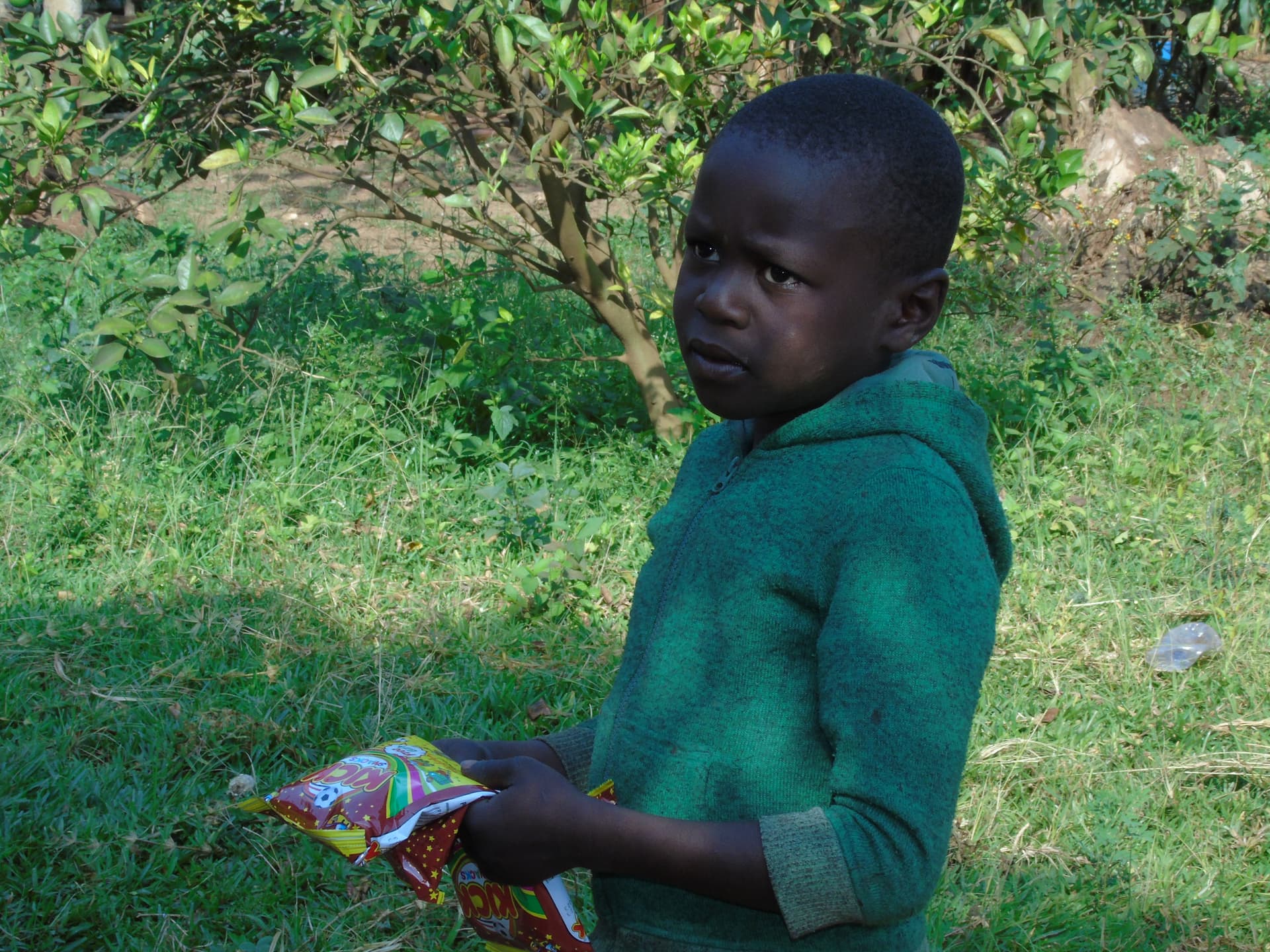 Boy in a green hoodie standing outdoors holding a colorful snack package during food and tract distribution.