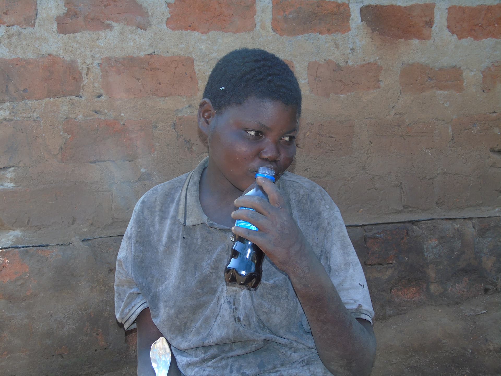 Boy in a gray polo shirt sitting against a brick wall, drinking from a bottle with a straw.