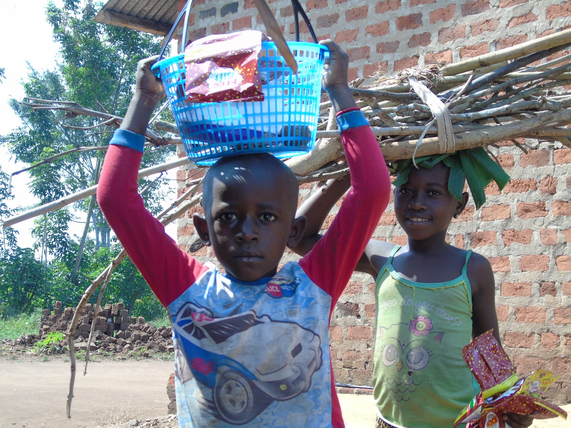 Two boys standing outdoors near a brick building, one carrying a blue plaid bag on his head.