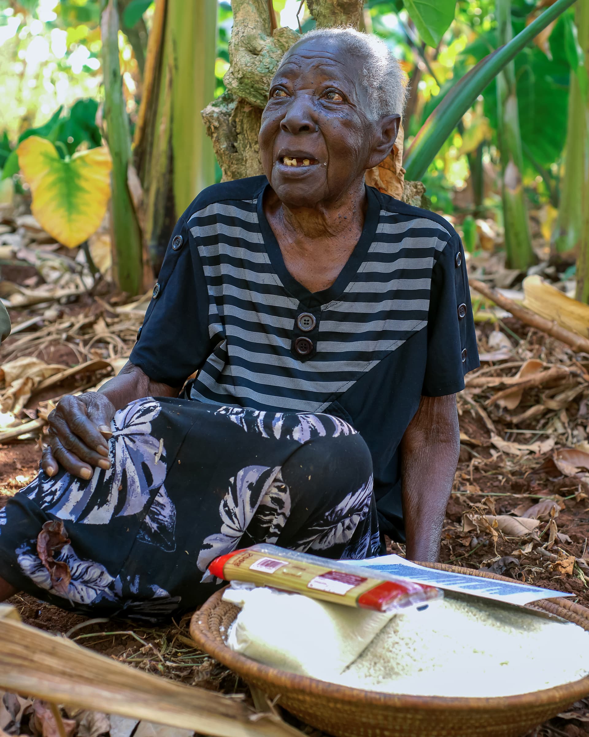 Elderly woman sits on the ground among banana leaves with a basin of food and gospel tracts.