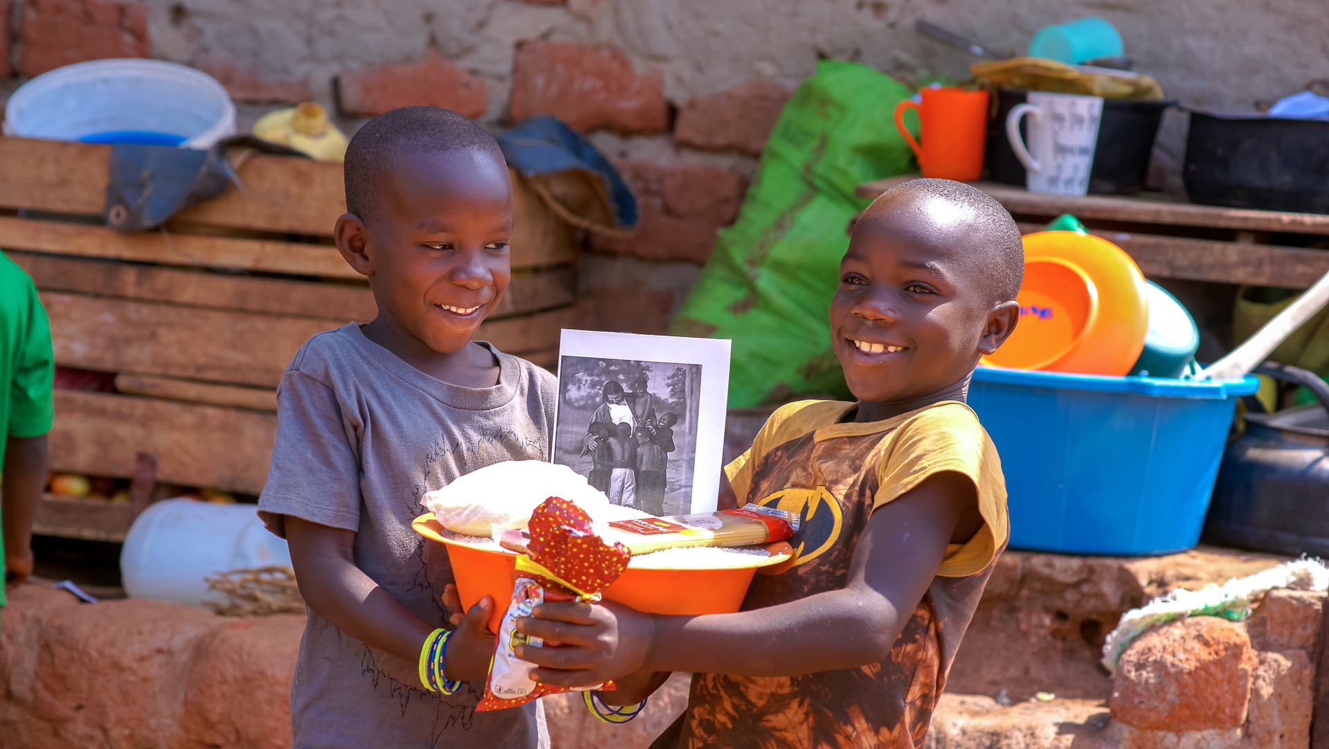 Two children hold a basin of food gifts, colorful plastic buckets in the background.