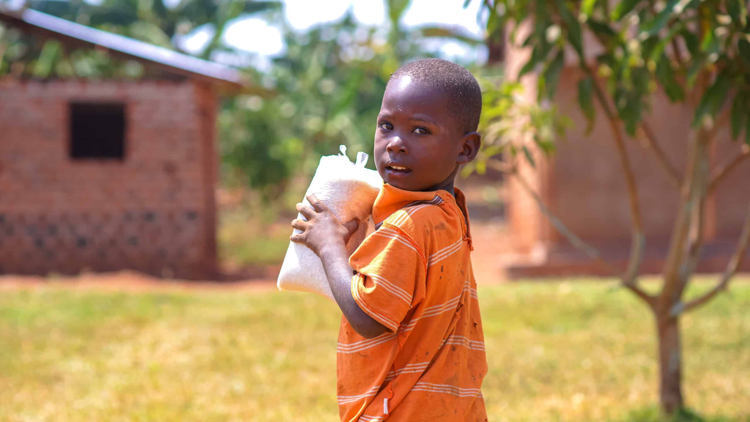 Young boy in an orange shirt drinks from a cup outdoors near a brick building.